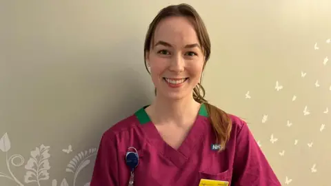 Ollie Conopo/BBC Woman with long brown hair smiling wearing a purple top with green around the neck.
