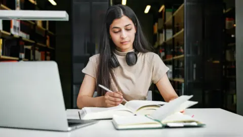 Tom Werner/Getty Images A woman sits in a university library with a laptop and two books, making notes