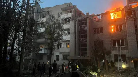 Firefighters work at a site of a building damaged by a Russian missile strike, amid Russia's attack on Lviv, Ukraine. A building is damaged and on fire, with firefighters standing in the foreground. 