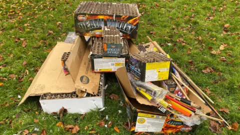 Boxes of used fireworks are left on grass and fallen leaves in a park.