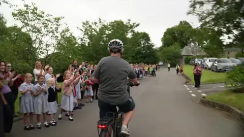 BBC/Mark Graham A line of children and parents is along the side of a long driveway, with a woman cycling down the middle, away from the camera.