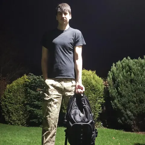 Kacper Bilengrek A young man with short brown hair standing in a garden at night. He is wearing a black T-shirt and sand-coloured combat trousers. He is holding a black rucksack. Green grass and shrubs can be seen beneath the black sky.