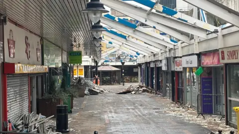 A derelict shopping centre with debris on the floor and shop window frames which have been ripped off.