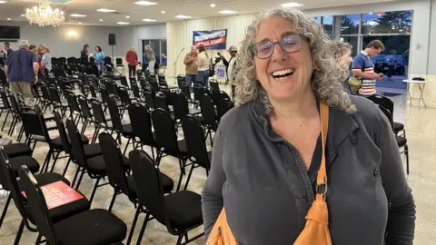 BBC/Anthony Zurcher A woman smiles in an event hall filled with chairs. A sign behind her says "Graham Platner"