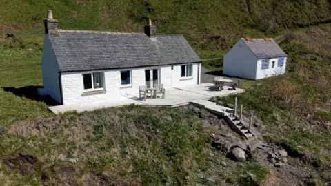 Galbraith A coastal cottage with whitewashed walls and decking in front and a wooden stairway leading down to the shore. There is a small hut beside it.