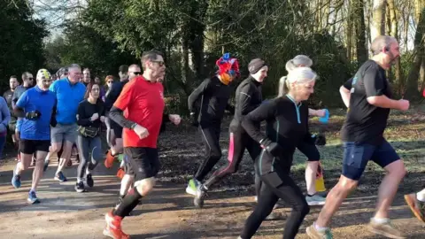 Around 20 runners in mostly black, blue and red sports gear jogging on a path in a forest setting.