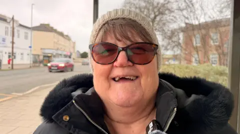 A woman smiling at the camera while standing outside on a path. She is wearing tortoise-shell sunglasses, a beige woolly hat and a black coat with a fluffy hood. Large buildings can be seen on either side of a road, with a red car driving past.