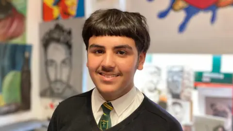 A teenager of South Asian heritage smiles toward the camera, he is wearing a dark green tie, white shirt and black jumper, behind him are pieces of hand drawn artwork on the wall.