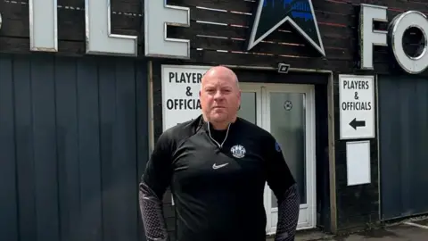 Gareth Edwards stands looking at the camera in front of the football club entrance. He has short white hair and is wearing the Newcastle Blue Star FC kit.