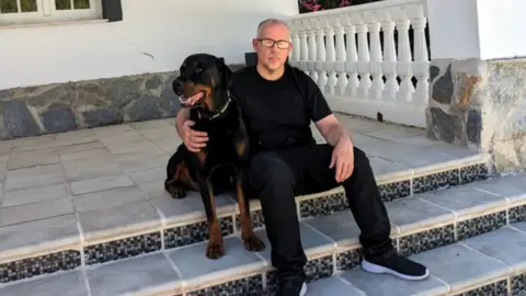 Ryan O'Meara Ryan is sitting on some steps outside a building with his right arm around his dog that's also sitting on the steps. He is dressed in a black t-shirt, black trousers and black trainers, with grey cropped hair and black glasses