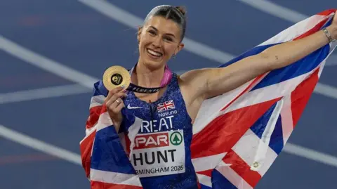 Amy Hunt - a woman in a sparkly blue Nike vest - is draped in a union jack flag which she is holding out into the air with one hand. In her other hand she is holding a gold circular medal. She has brown hair slicked back and is smiling. A blurred racetrack can be seen behind her.