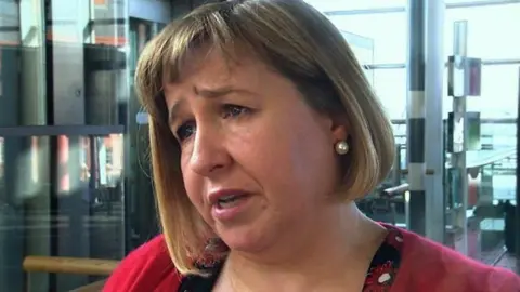 Lynne Neagle, with a red top, in front of part of the Senedd. It is a head and shoulders shot of her.