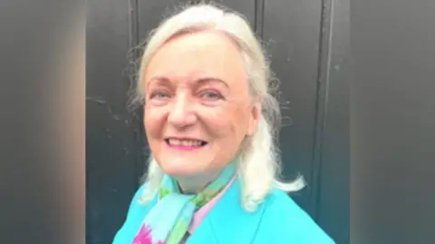 A woman with blond shoulder length hair and pink lipstick smiling . She is turned towards the camera but her shoulders are angled away. The shoulders of her turquoise jacket can e seen and she is wearing a turquoise, pink and green neck scarf. A dark grey fence can be seen behind her.