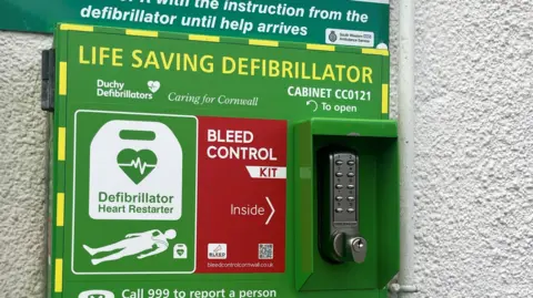 A green cabinet on a white wall with a security handle to open, it is covered in lettering which explains it is a life saving defibrillator from Duchy Defibrillators. It also has a red sticker on it which says bleed control kit inside.