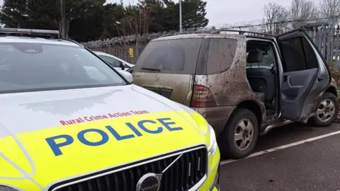A yellow and white marked police care is in the foreground, with RURAL CRIME ACTION TEAM printed in red on the bonnet. Behind is a 4x4 vehicle which is splattered with mud.