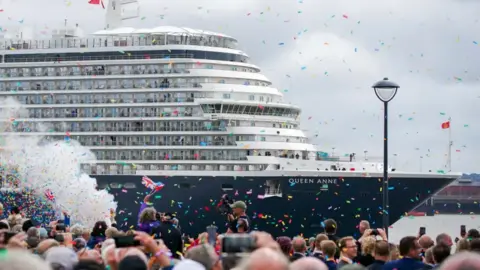 PA Media A wide shot of the Queen Anne cruise ship arriving into the Mersey. It is an overcast day, but hundreds of spectators are celebrating with colourful confetti.