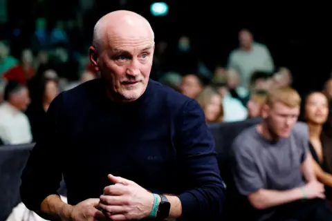 PA Media Barry McGuigan watches a boxing match at ringside. He has a bald head, a grey beard and is wearing a blue jumper with the sleeves rolled up. A large crowd is visible behind him.