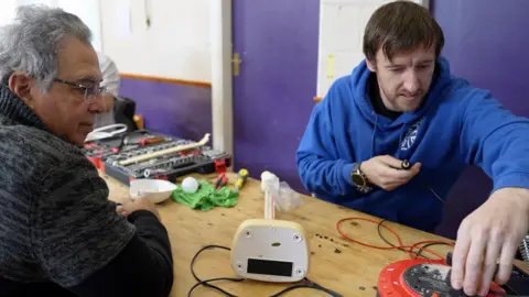 A man with glasses and wearing a grey jumper looks on as a man in a blue hoodie puts a plug into an extension socket with one hand while holding a soldering iron in the other.