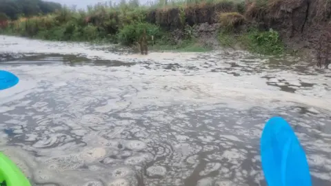 Ben James Picture of the river Tywi banks with a yellow discharge floating on the surface. There are two blue oars on the edge of the frame and the edge of a green kayak.