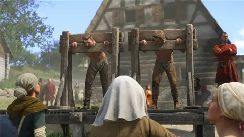 Warhorse Studios A screenshot shows two characters locked inside stocks in the centre of a Medieval town square. They are on a platform surround by a crowd of peasants.