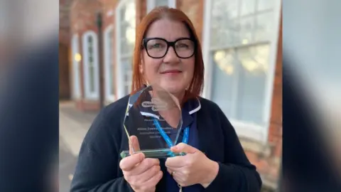 A woman with red hair and black-rimmed glasses smiling at the camera. She is holding a clear trophy and wearing a navy blue nurse's top. There is a brick building behind her.