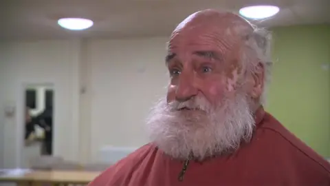 Head and shoulders image of Alan Jones. He has a bald head with longer white hair at the sides and a long beard. He is wearing a red jacket