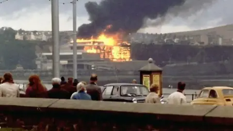 An old photograph of people looking on at the complex, which is on fire at the other side of Douglas Bay.