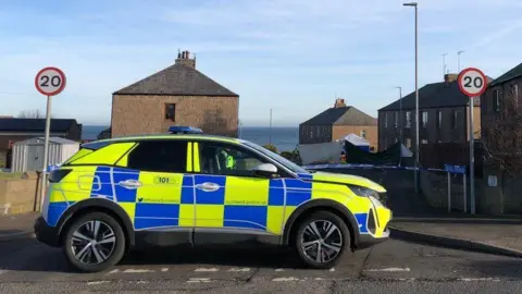 Police car with yellow and blue markings at the scene of an accident, with caution tape in the background and two 20 mph speed limit signs and a blue sky.