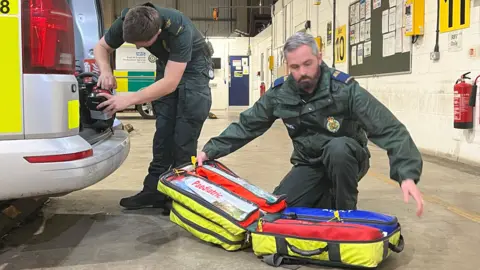 Two co-responders pack an ambulance van, one at the boot, another one kneels on the floor with equipment
