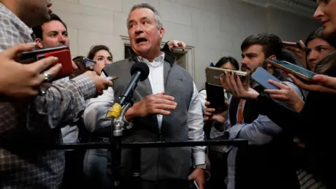 Getty Images Don Bacon, wearing a grey sweater vest over a white dress shirt, gestures as he answers questions surrounded by a gaggle of Capitol Hill reporters aiming microphones and phones in his direction