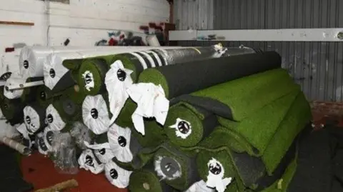 NCA A stack of rolled, artificial grass in a warehouse.