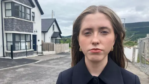 Sinn Féin councillor Áine Quinn standing in front of the two-storey house in Sliabh Mor where a pipe bomb exploded overnight. Quinn has long, straight fair hair and is wearing a black coat with a large collar and gold earrings.