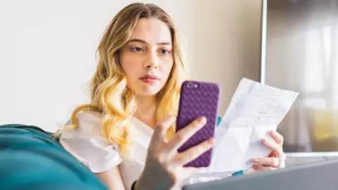 Getty Images Una joven rubia con expresión concentrada mira fijamente su teléfono inteligente mientras sostiene un billete en la otra mano.