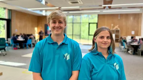 A young man and a young woman are standing in a large classroom, wearing turquoise T-shirts. The man in on the left and has short blond hair, whilre the woman has shoulder length dark blond hair with blue highlights.