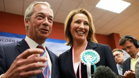 Nigel Farage, in a dark blue suit, and Sarah Pochin, in a blazer wearing a blue and white Reform UK rosette, beam at the camera.