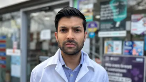 Matt Hutchinson / BBC A young man with short hair and a beard is wearing a white coat. He is looking stern and standing in front of a shop front. 