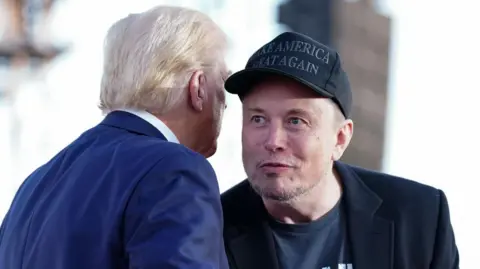 Getty Images Tesla and SpaceX CEO Elon Musk speaks with former president Donald Trump during a campaign event in Pennsylvania. Musk wears a black 'Make America Great Again' hat and black blazer while Trump wears a blue suit.