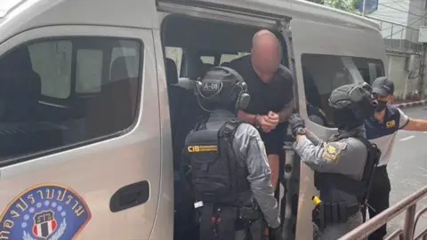 Essex Police A man with his face blurred walking out of a Thailand Police car van with two Thai Police officers standing in front of him. The man is wearing a black top with his hands in front of him.