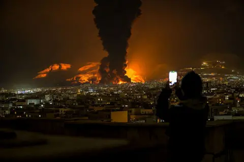 Getty Images A woman uses her phone to record as smoke and flames rise at the site of airstrikes on an oil depot in Tehran on 7 March, 2026. 
