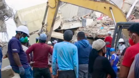 A crowd in hard hats and caps gathers around rubble with an excavator in the middle.