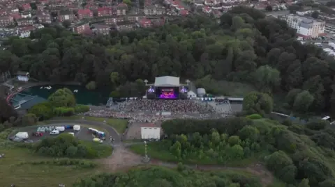 Cuffe and Taylor An aerial picture of Scarborough's open air theatre, the stage is lit up in thw centre, with a large crowd in front, it's surrounded by trees and buildings.
