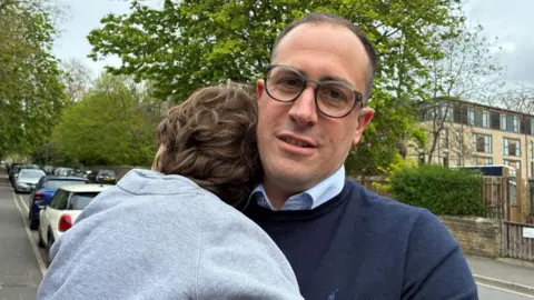 A man stands in a leafy street, with cars parked on both sides of the road behind him. He is looking into the camera and is wearing glasses with dark frames. He is wearing a dark blue jumper over a blue shirt. He is holding a child with dark hair who is wearing a grey top.