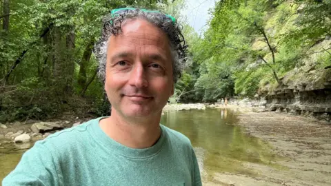 Handout A man in a green sweatshirt with short black and grey hair smiling in front of a lake
