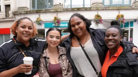 Serena Hancock is stood to the left holding a takeaway hot drink cup, she is wearing a black and white sports jacket and has her hair styled in cornrows and a ponytail, Chloe Griffiths is wearing a brown and beige psychedelic jacket and a matching brown top. Her brown hair is pulled back in a ponytail, Tianna Hancock has her arm around Chloe and Amaris who is stood next to her. Tianna is wearing a black jacket and a white knitted jumper. Her brown curly hair is short and loose, and Amaris Powell is wearing a black rain jacket and a red 3/4 zip jumper. Her black hair is pulled into a bun