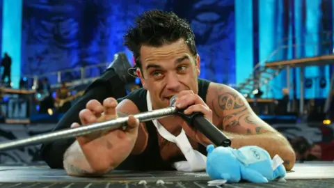 Getty Images A young Robbie Williams is laying on his front on a stage with dramatic blue lighting behind him illuminating his band and stage, he is holding a microphone to his mouth