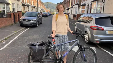 Meghan Hoyt Meghan Hoyt standing next to her bike on a residential street. She has blonde curly hair, wearing sunglasses on her head, a cream jumper, clue floral skirt and yellow backpack.