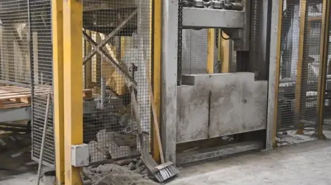 HSE A concrete weight can be seen exposed in a factory environment. There is lots of concrete dust around the site and a broom and a spade. Other parts of the machinery and site can be seen to be closed off behind metal fencing. 