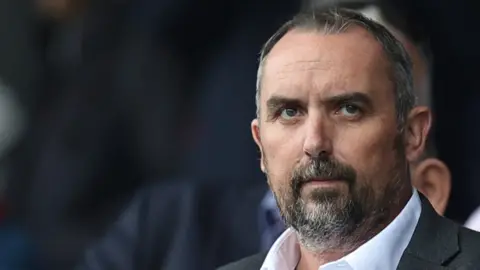 Close-up of Kelvin Thomas standing in a white open-necked shirt and black blazer in a football crowd. He has dark/grey hair.