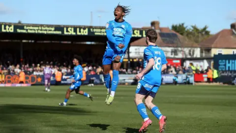 Getty Images Tayo Edun of Peterborough United jumping in the air in front of another Posh player and punching the air. He appears to be shouting in celebration. Both are wearing light blue kits. In the background fans watch on from the stands. 