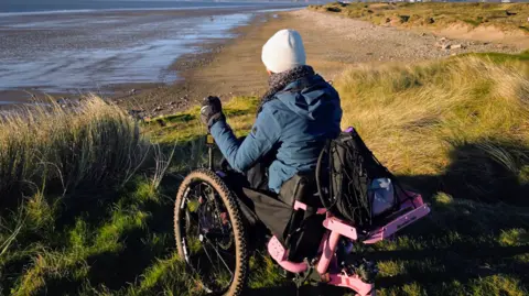 Bethany Handley Bethany in a motorised all-terrain trike wheelchair, which has a pink metal structure supporting a back wheel. She is photographed from behind wearing a blue coat, cream hat and grey scarf and gloves, looking down from the common over Sker Beach towards Swansea Bay.